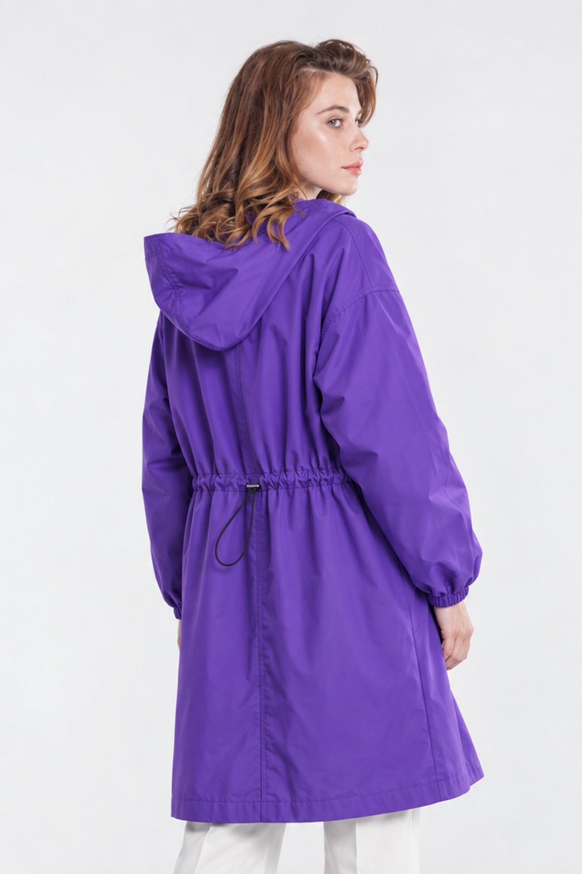 A woman with wavy brown hair wearing the Icon Milano Windbreaker Rain Jacket—a long, purple coat with a cinched waist and hood—stands with her back to the camera, glancing over her shoulder. This stylish jacket boasts a water-resistant finish, while the plain light-colored background highlights its sleek silhouette.