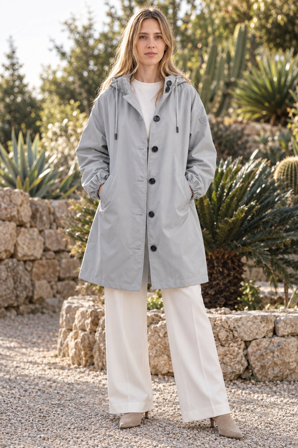 A woman stands outdoors on a gravel path, her hands in the pockets of the light gray Icon Milano Windbreaker Rain Jacket layered over a white shirt and wide-leg white pants. Stone walls and green plants provide a lush backdrop.