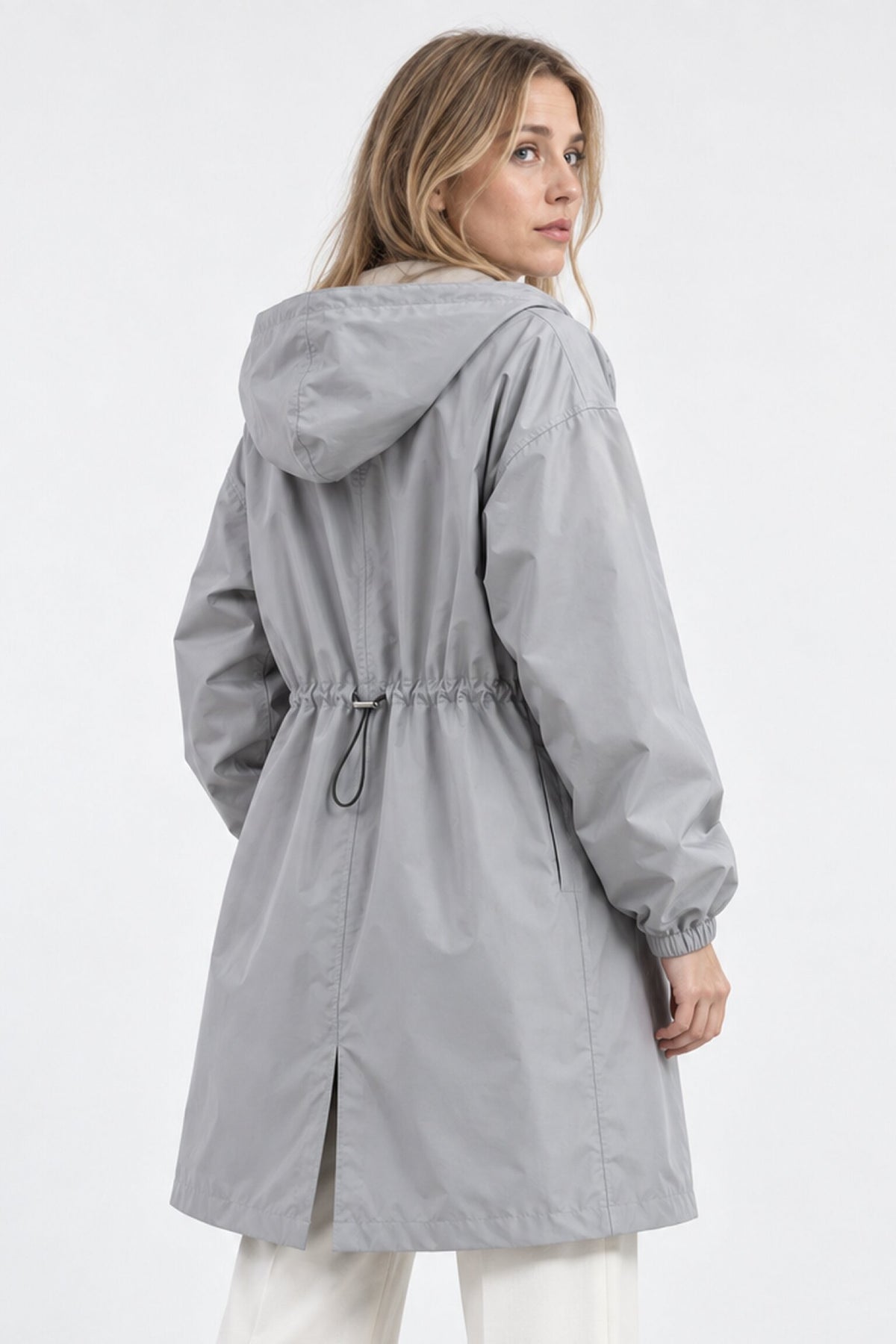 A woman with long blonde hair wears the Icon Milano Windbreaker Rain Jacket in light gray, a windproof outerwear piece featuring a drawstring waist and back vent. The longline rain jacket is styled facing mostly away from the camera against a plain background.