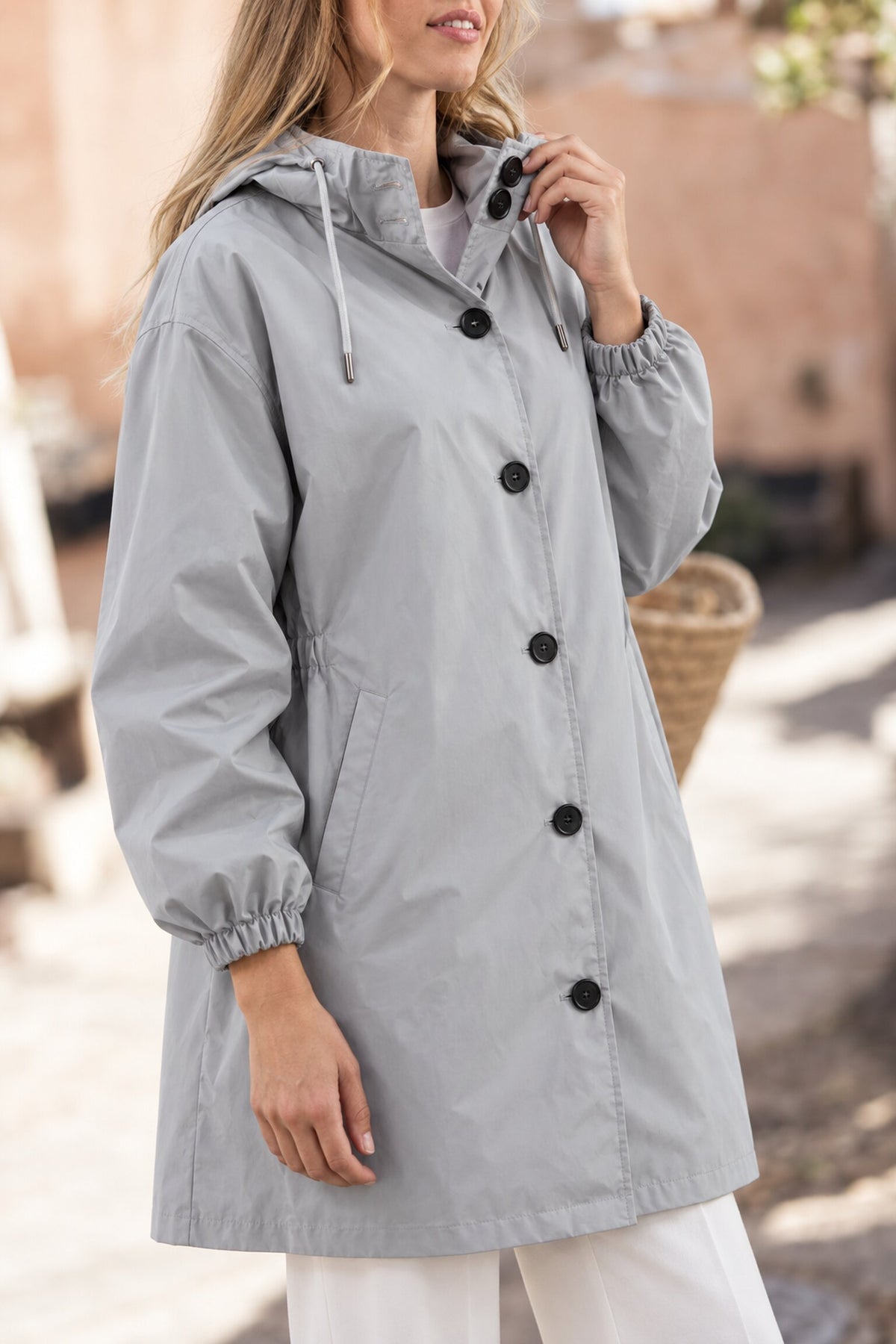 A person stands outdoors wearing the Icon Milano Windbreaker Rain Jacket—a light gray, longline coat featuring black buttons, a hood, and elastic cuffs. The blurred background reveals hints of beige tones and natural light. This windproof outerwear combines style with reliable protection for uncertain weather conditions.