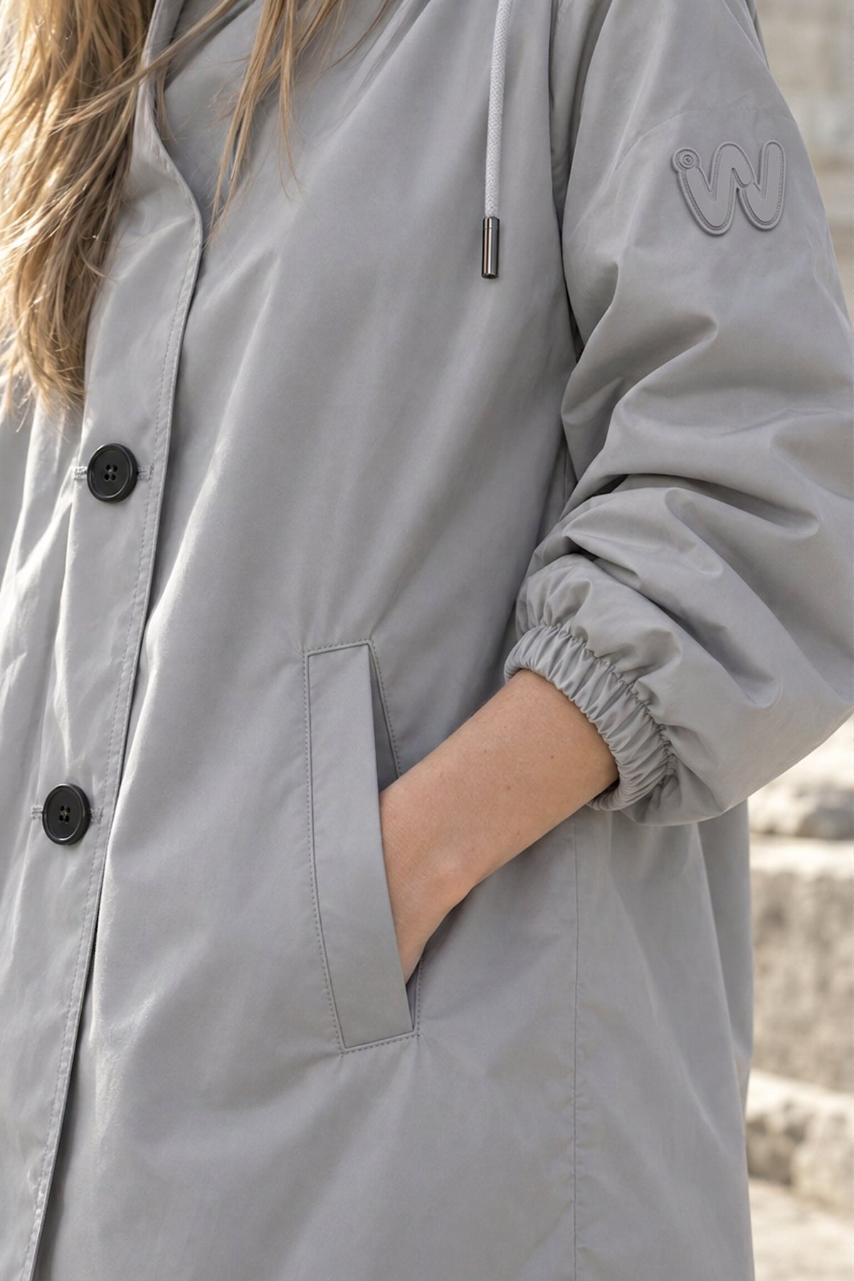 A person with long, light brown hair wears the Icon Milano Windbreaker Rain Jacket in light gray, featuring black buttons and a logo on the sleeve. One hand is tucked into the jacket pocket, and stone steps form the background.