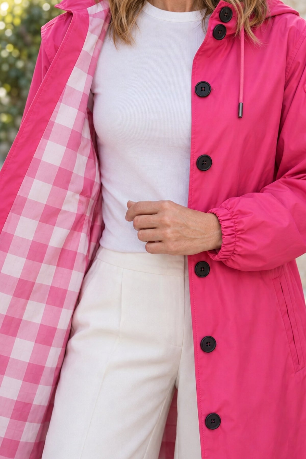 A person models the Icon Milano Windbreaker Rain Jacket, an Italian-designed women’s outerwear coat in hot pink with black buttons, revealing its pink and white checkered lining. They are also dressed in a white shirt and cream pants. The photo is cropped above the nose.