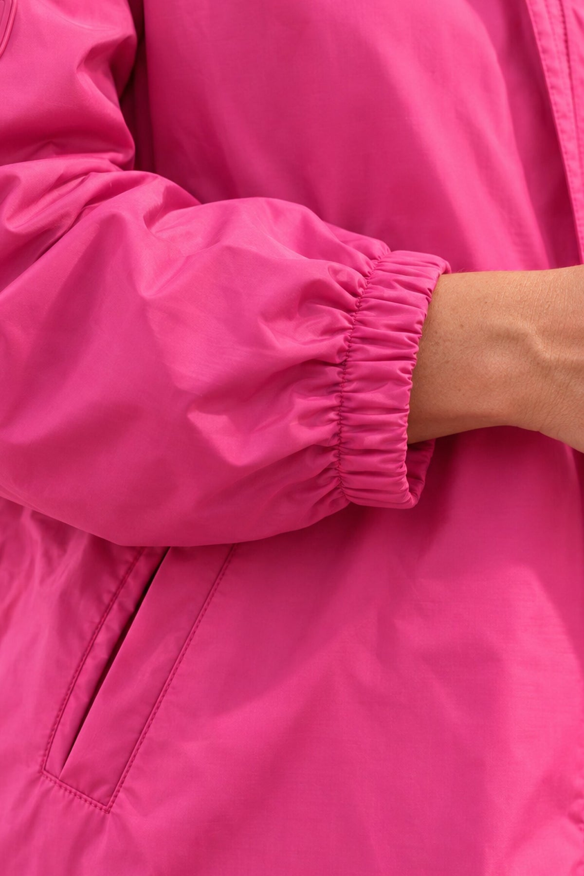 A close-up of a person wearing the Icon Milano Windbreaker Rain Jacket—a bright pink, Italian-designed women’s outerwear piece with an elastic cuff. Their hand is partially visible, and the water-resistant jacket features a side pocket and a visible sleeve seam.