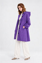A person stands sideways wearing the Icon Milano Windbreaker Rain Jacket—a long, purple, water-resistant hooded coat—over a light turtleneck and white pants, paired with beige heeled shoes, against a plain light background.