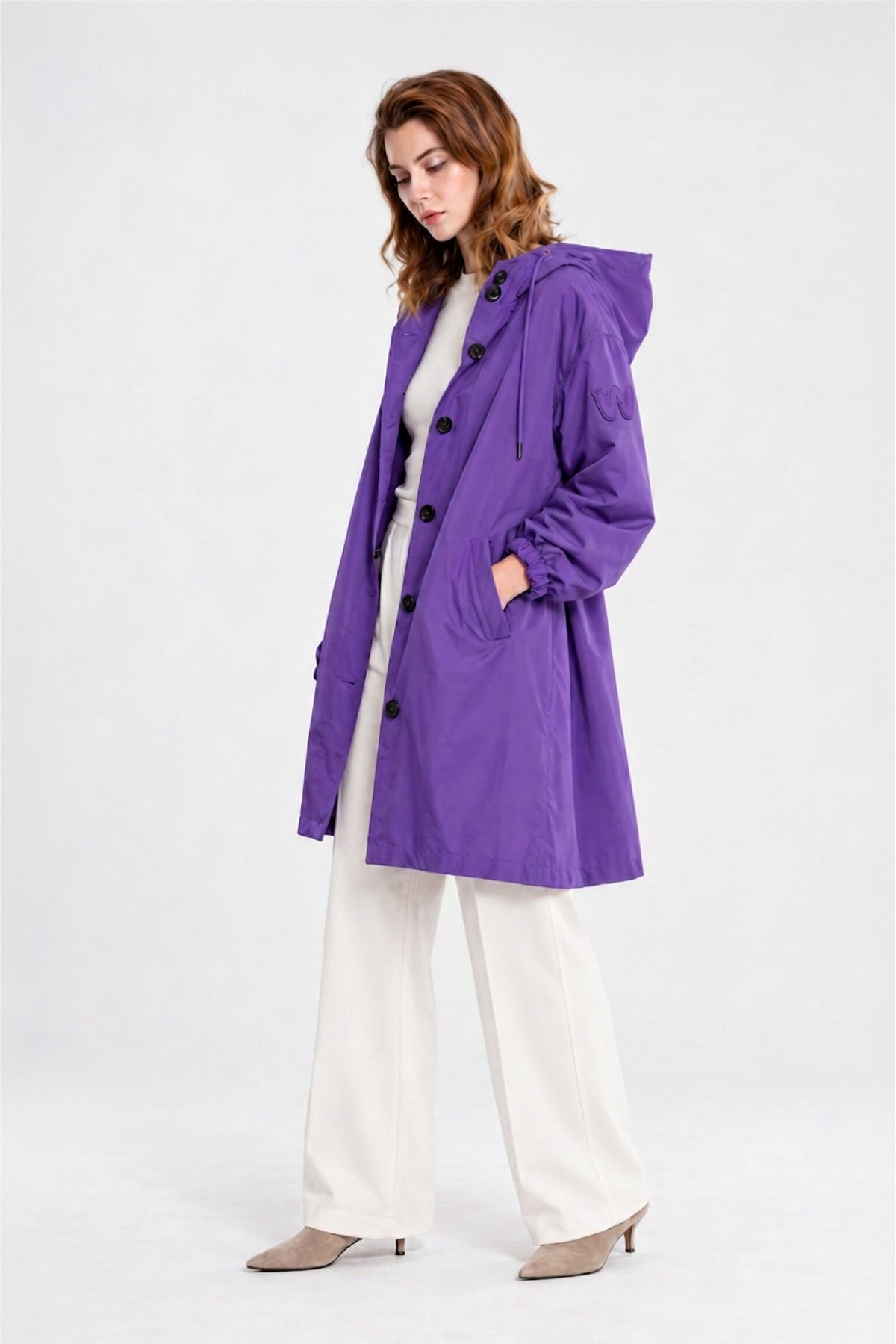 A person stands sideways wearing the Icon Milano Windbreaker Rain Jacket—a long, purple, water-resistant hooded coat—over a light turtleneck and white pants, paired with beige heeled shoes, against a plain light background.