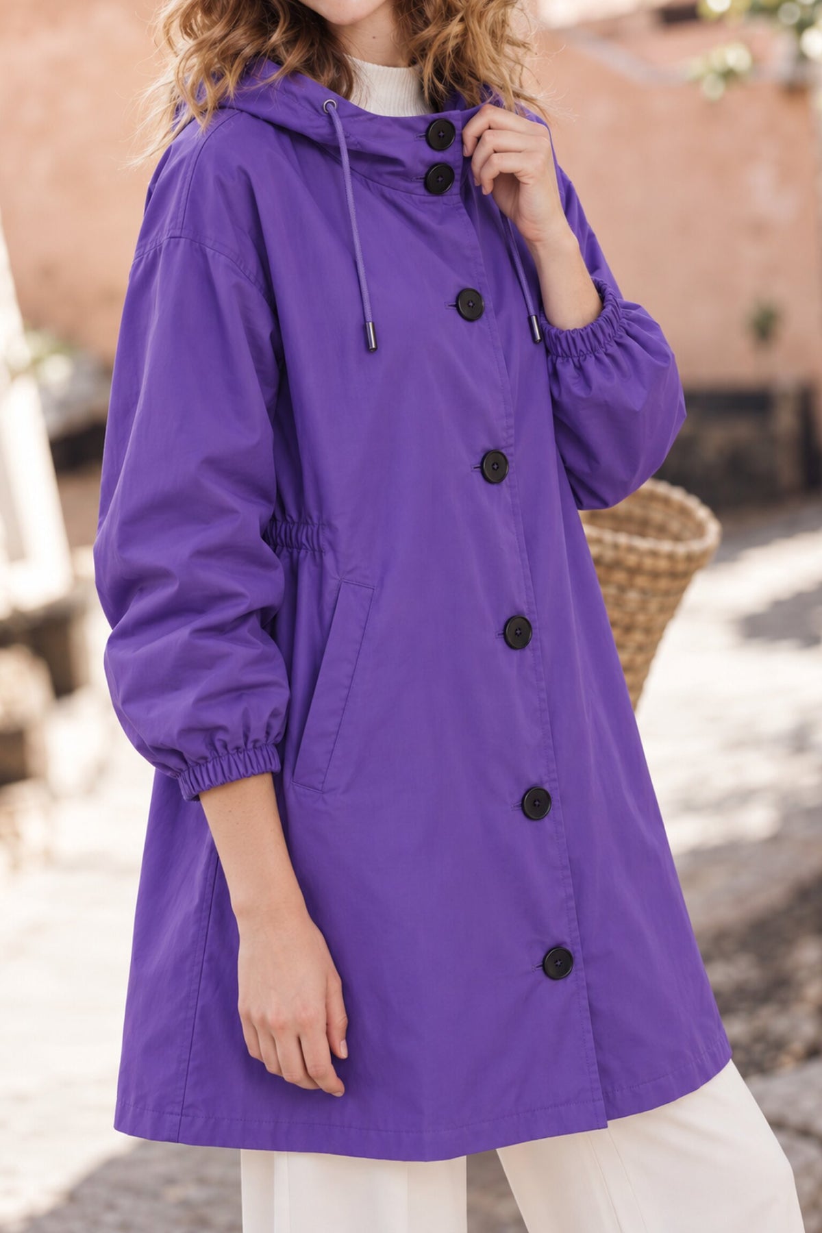 A person wearing an Icon Milano Windbreaker Rain Jacket in purple with black buttons and white pants stands outdoors, holding a woven bag. Their face is not visible in the image.