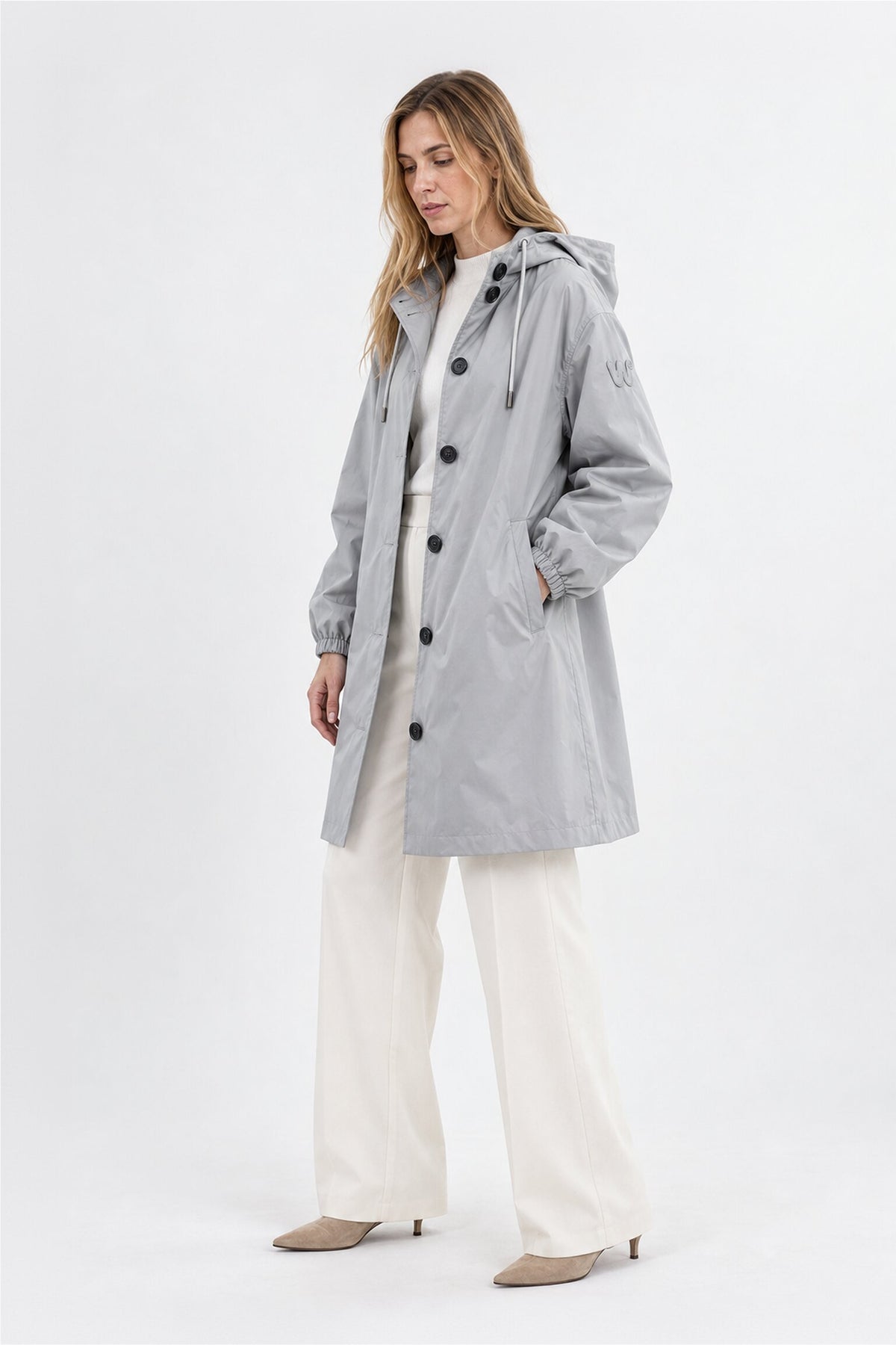 A woman stands wearing the Icon Milano Windbreaker Rain Jacket in light gray, featuring black buttons and a practical hood over a white shirt and wide-leg white pants. She has light brown hair and is styled with beige heeled shoes. The longline, water-resistant jacket blends practicality with style, set against a plain white background.