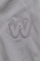 A gray fabric background featuring an embroidered, stylized “W” logo outlined in white captures the modern aesthetic of the Icon Milano Windbreaker Rain Jacket. A small registered trademark symbol is situated near the top left of the “W.”.
