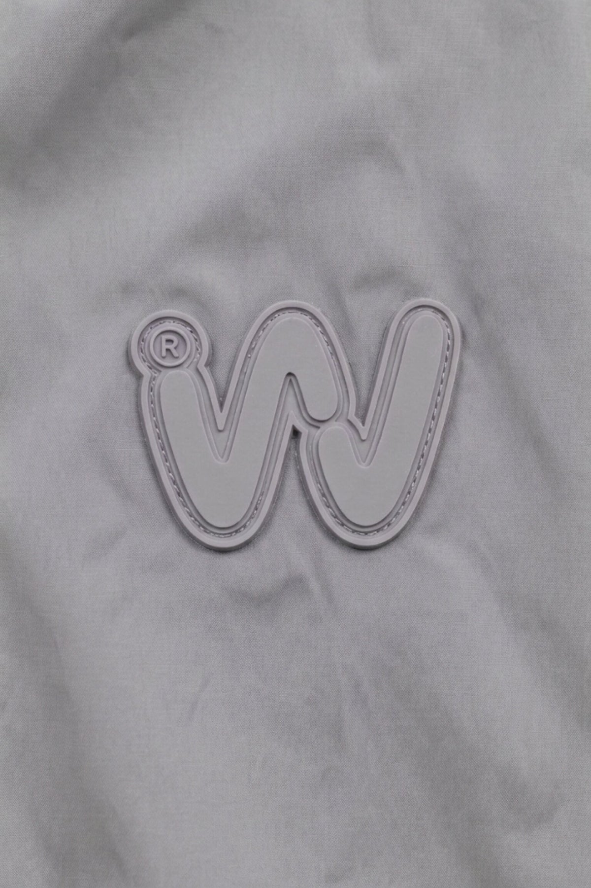 A gray fabric background featuring an embroidered, stylized “W” logo outlined in white captures the modern aesthetic of the Icon Milano Windbreaker Rain Jacket. A small registered trademark symbol is situated near the top left of the “W.”.