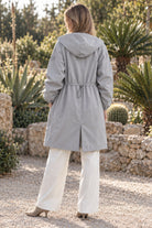 A person with light brown hair, dressed in the Icon Milano Windbreaker Rain Jacket in light gray and white pants, stands outdoors on a gravel path with their back to the camera, surrounded by stone walls, lush green plants, and cacti.