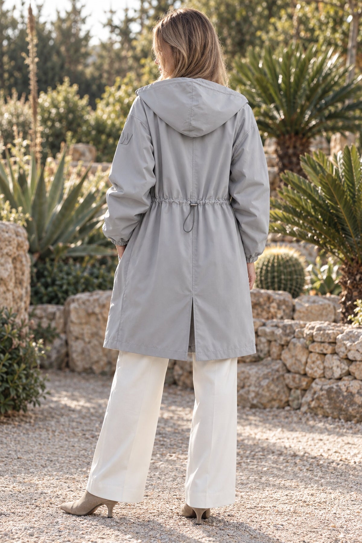 A person with light brown hair, dressed in the Icon Milano Windbreaker Rain Jacket in light gray and white pants, stands outdoors on a gravel path with their back to the camera, surrounded by stone walls, lush green plants, and cacti.