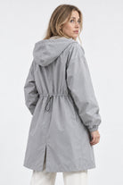 A woman with long blonde hair wears the Icon Milano Windbreaker Rain Jacket in light gray, a windproof outerwear piece featuring a drawstring waist and back vent. The longline rain jacket is styled facing mostly away from the camera against a plain background.