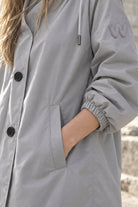 A person with long, light brown hair wears the Icon Milano Windbreaker Rain Jacket in light gray, featuring black buttons and a logo on the sleeve. One hand is tucked into the jacket pocket, and stone steps form the background.