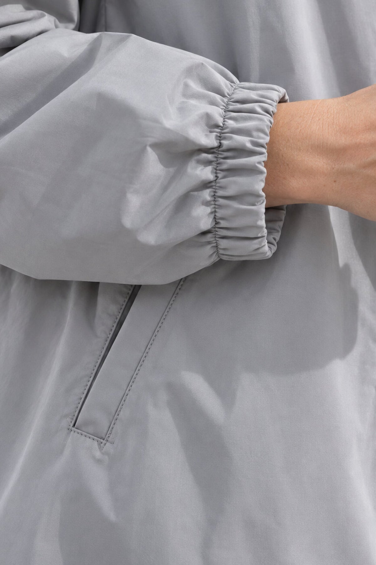 A close-up of a person’s arm wearing the Icon Milano Windbreaker Rain Jacket in light gray, a water-resistant women’s coat with elastic at the wrist. The longline rain jacket features a visible pocket, and the person’s hand is partially shown.