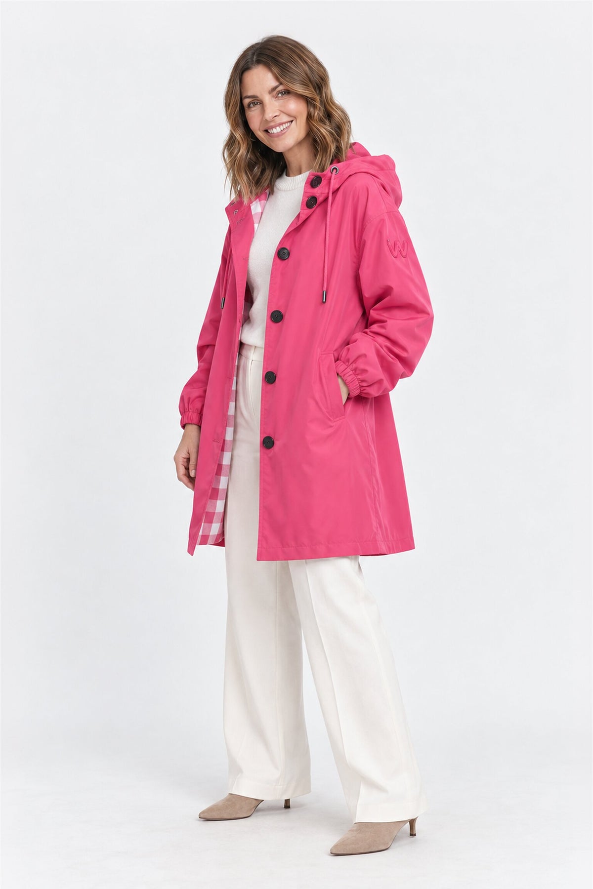 A woman stands smiling, wearing the Icon Milano Windbreaker Rain Jacket in bright pink with black buttons, layered over a white top and white pants. She has one hand in her pocket and is paired with beige heeled shoes against a plain white background.
