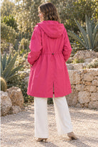 A woman stands outdoors on a gravel path, facing away, wearing the Icon Milano Windbreaker Rain Jacket in bright pink and white pants. Stone walls, green plants, and sunlight filtering through trees make up the background.