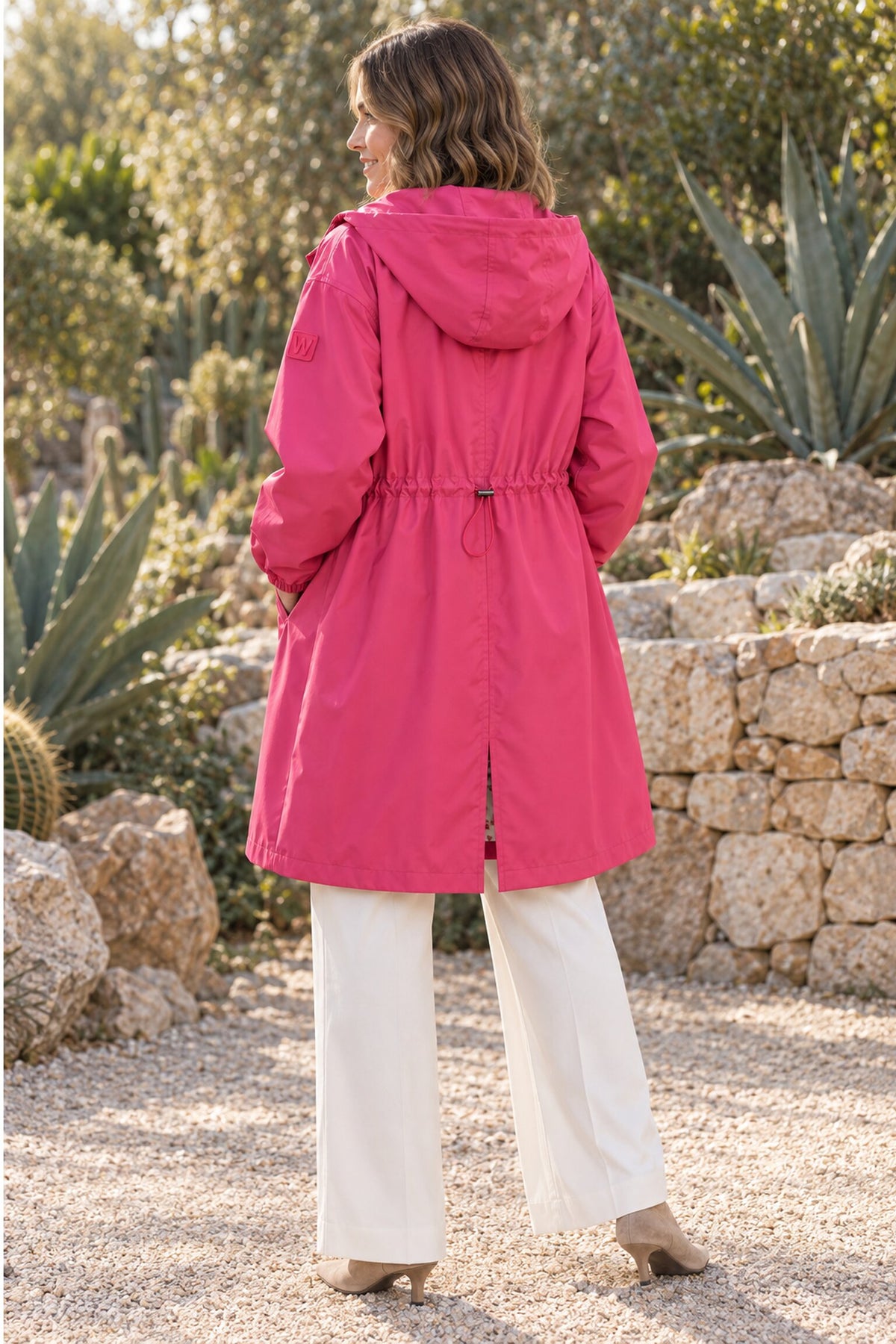 A woman stands outdoors on a gravel path, facing away, wearing the Icon Milano Windbreaker Rain Jacket in bright pink and white pants. Stone walls, green plants, and sunlight filtering through trees make up the background.