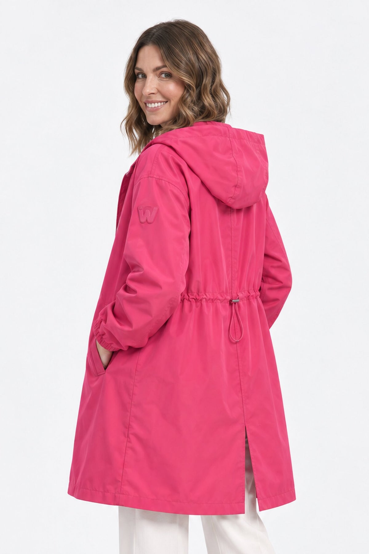 A woman with wavy brown hair is smiling as she turns to show the back of the bright pink Icon Milano Windbreaker Rain Jacket she’s wearing. This longline, water-resistant coat features a drawstring waist, side pockets, a small slit at the hem, and a “W” logo on the sleeve.