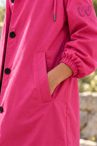 A person wears the Icon Milano Windbreaker Rain Jacket, a bright pink, Italian-designed women’s outerwear coat with black buttons, and has their right hand in the pocket. Only the torso and arm are visible against a blurred outdoor background. The sleeve of this water-resistant women’s jacket features a small embroidered logo near the shoulder.