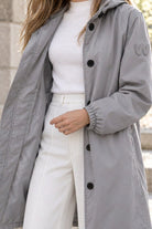 A person stands outdoors wearing the Icon Milano Windbreaker Rain Jacket—a long, light gray, water-resistant women’s coat with a patch on the upper sleeve—layered over a white turtleneck and white pants. The person’s head is not visible in the image.