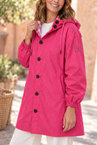 Wearing the Icon Milano Windbreaker Rain Jacket in vibrant pink with black buttons and front pockets, a woman models outdoors beside a wicker basket, surrounded by gentle greenery and pale walls.