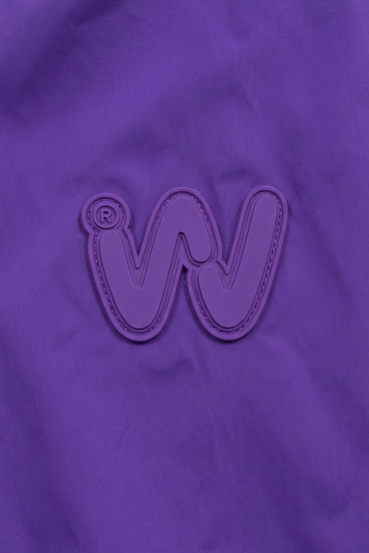 A raised, stylized “W” logo with a registered trademark symbol appears on a solid purple fabric background, evoking the signature look of the Icon Milano Windbreaker Rain Jacket.