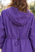A person with wavy, light brown hair is wearing a purple Icon Milano Windbreaker Rain Jacket with a cinched waist, viewed from the back. The background is outdoors and slightly blurred.