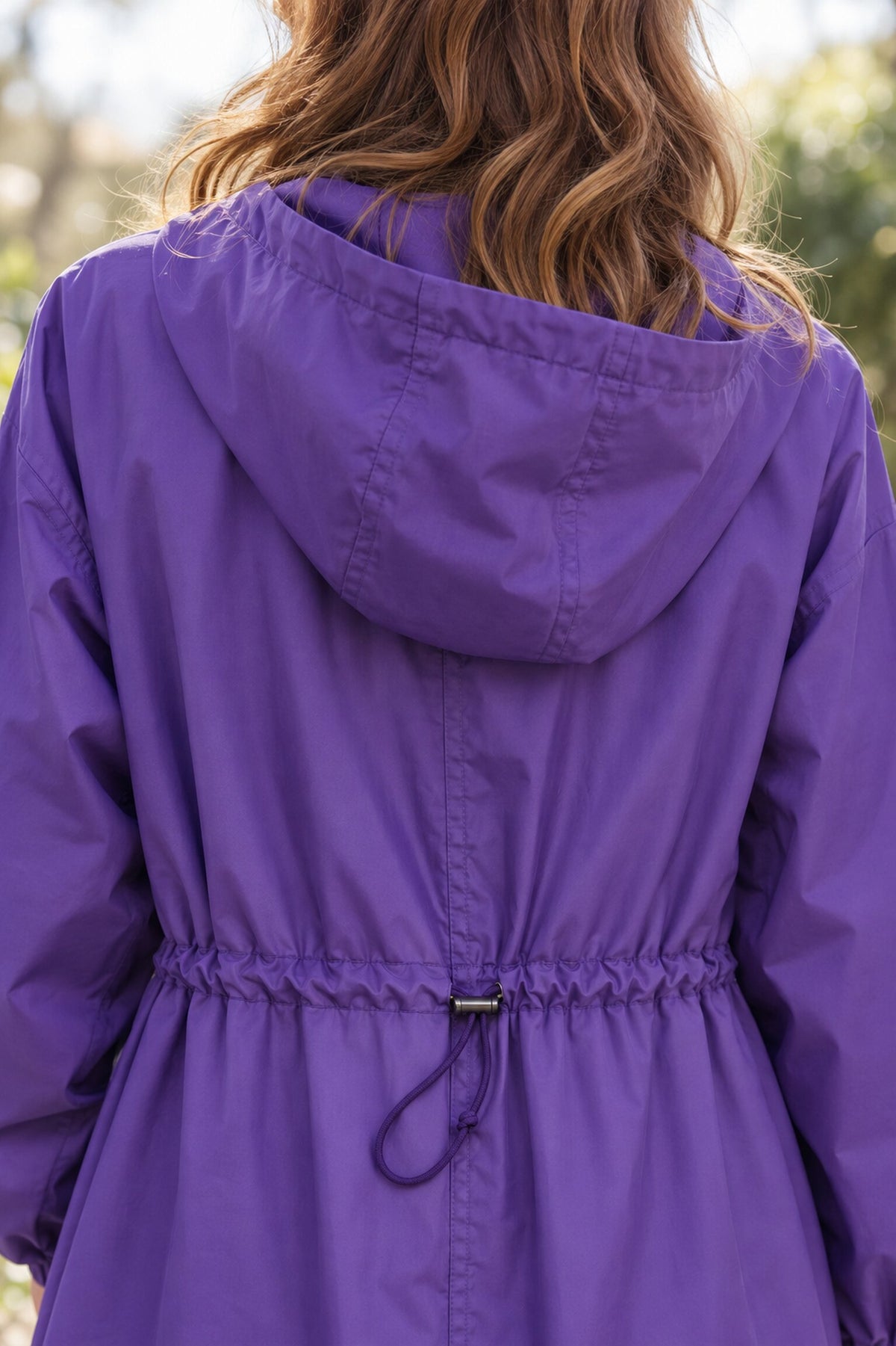 A person with wavy, light brown hair is wearing a purple Icon Milano Windbreaker Rain Jacket with a cinched waist, viewed from the back. The background is outdoors and slightly blurred.