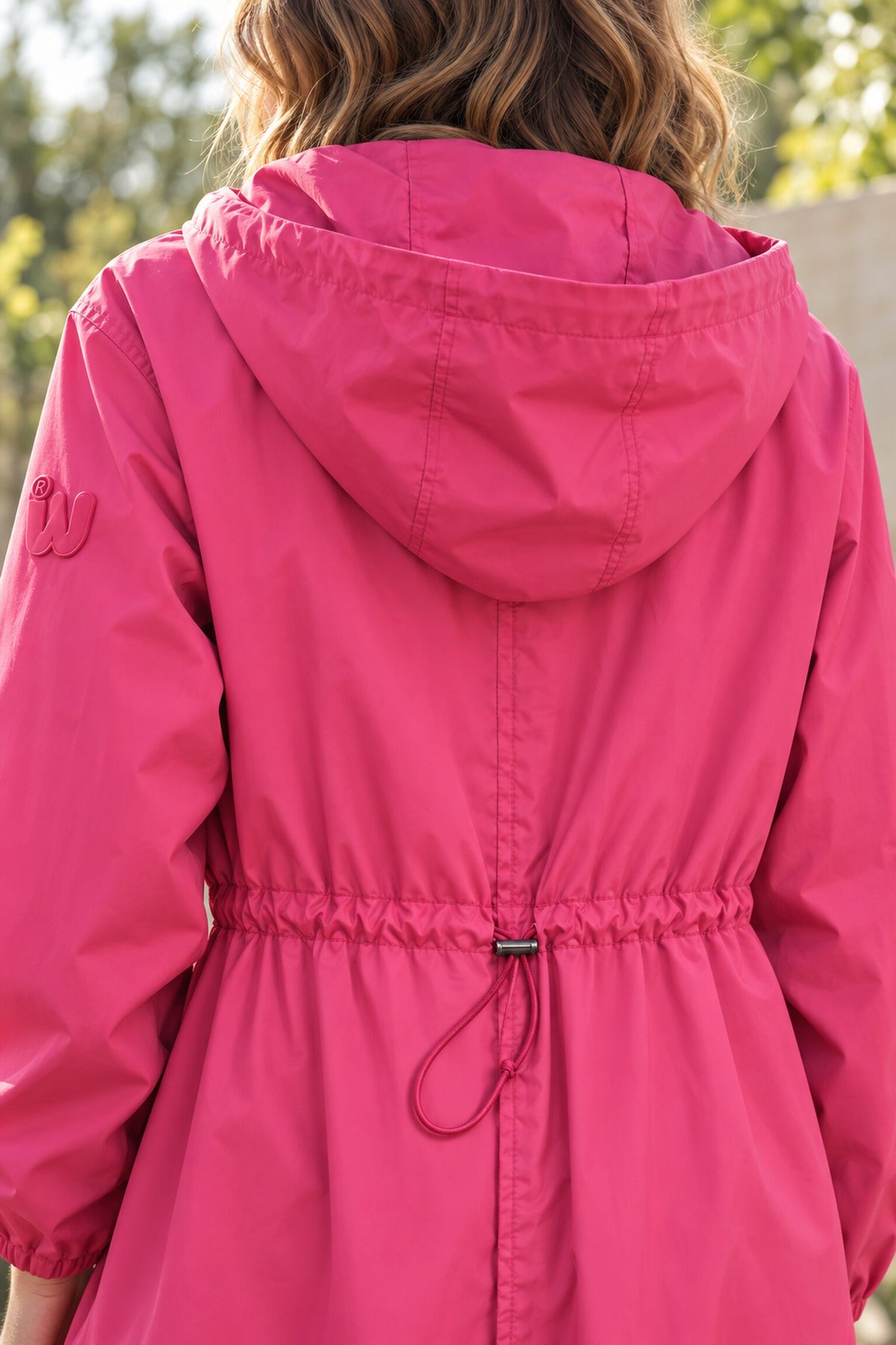 A person with wavy hair is pictured from behind wearing the bright pink Icon Milano Windbreaker Rain Jacket for women, featuring an adjustable drawstring at the waist. Green trees and sunlight are visible in the blurred background.