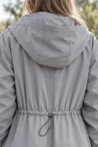 A person is shown from the back wearing the Icon Milano Windbreaker Rain Jacket in light gray. This windproof longline outerwear features an adjustable drawstring at the waist and is worn outdoors, with greenery visible in the background.