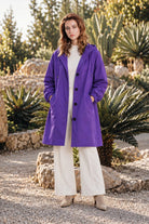 A woman stands outdoors on gravel, wearing the Icon Milano Windbreaker Rain Jacket—a long purple, water-resistant coat—layered over a cream sweater and wide-leg cream pants. She has light brown hair and beige ankle boots, with lush green plants and rocks in the background.