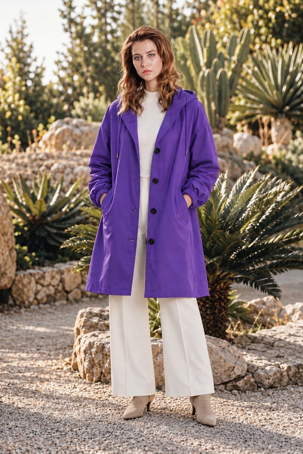 A woman stands outdoors on gravel, wearing the Icon Milano Windbreaker Rain Jacket—a long purple, water-resistant coat—layered over a cream sweater and wide-leg cream pants. She has light brown hair and beige ankle boots, with lush green plants and rocks in the background.