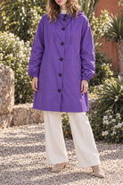 A person stands outdoors on a gravel path wearing the Icon Milano Windbreaker Rain Jacket—a long, purple, water-resistant hooded coat—paired with white wide-leg pants and beige shoes. The background features green plants and flowering bushes. The person’s face is partially visible.