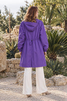 A person is standing outdoors on a gravel path, facing away. They are wearing the Icon Milano Windbreaker Rain Jacket in purple and white pants. Green plants and stone walls are visible in the background.