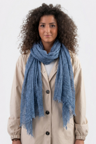 A person with curly hair models the Lightweight Linen Scarf—a textured, versatile accessory—paired with a beige buttoned coat, standing against a plain light background and facing the camera with a neutral expression.