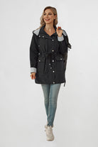 A woman stands against a plain white background, smiling and holding the strap of a black backpack on her shoulder. She is wearing a black STRIPED CARBON BLACK WINDBREAKER JACKET with a lightweight design from looselyboho, light blue jeans, and white sneakers. The jacket has a drawstring waist and silver buttons.