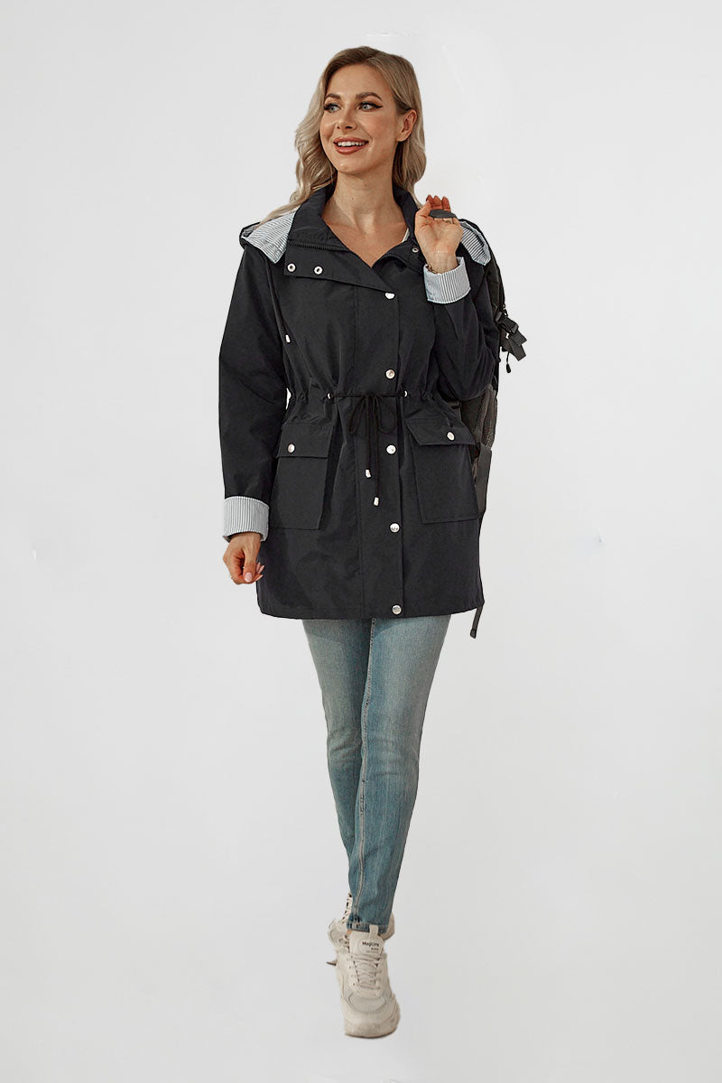 A woman stands against a plain white background, smiling and holding the strap of a black backpack on her shoulder. She is wearing a black STRIPED CARBON BLACK WINDBREAKER JACKET with a lightweight design from looselyboho, light blue jeans, and white sneakers. The jacket has a drawstring waist and silver buttons.